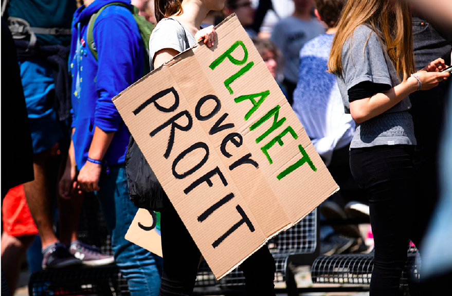 Photo by Markus Spiske on Unsplash. Protestor walking by with a sign that states "Planet over Profit."