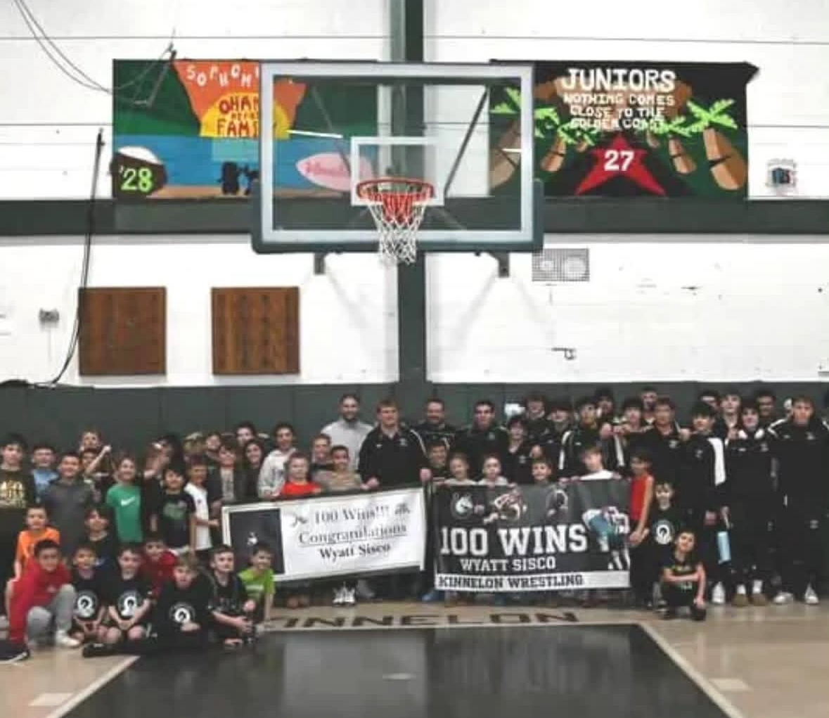 Wyatt Sisco along with recreational wrestlers taking a photo with his 100 wins banner. 